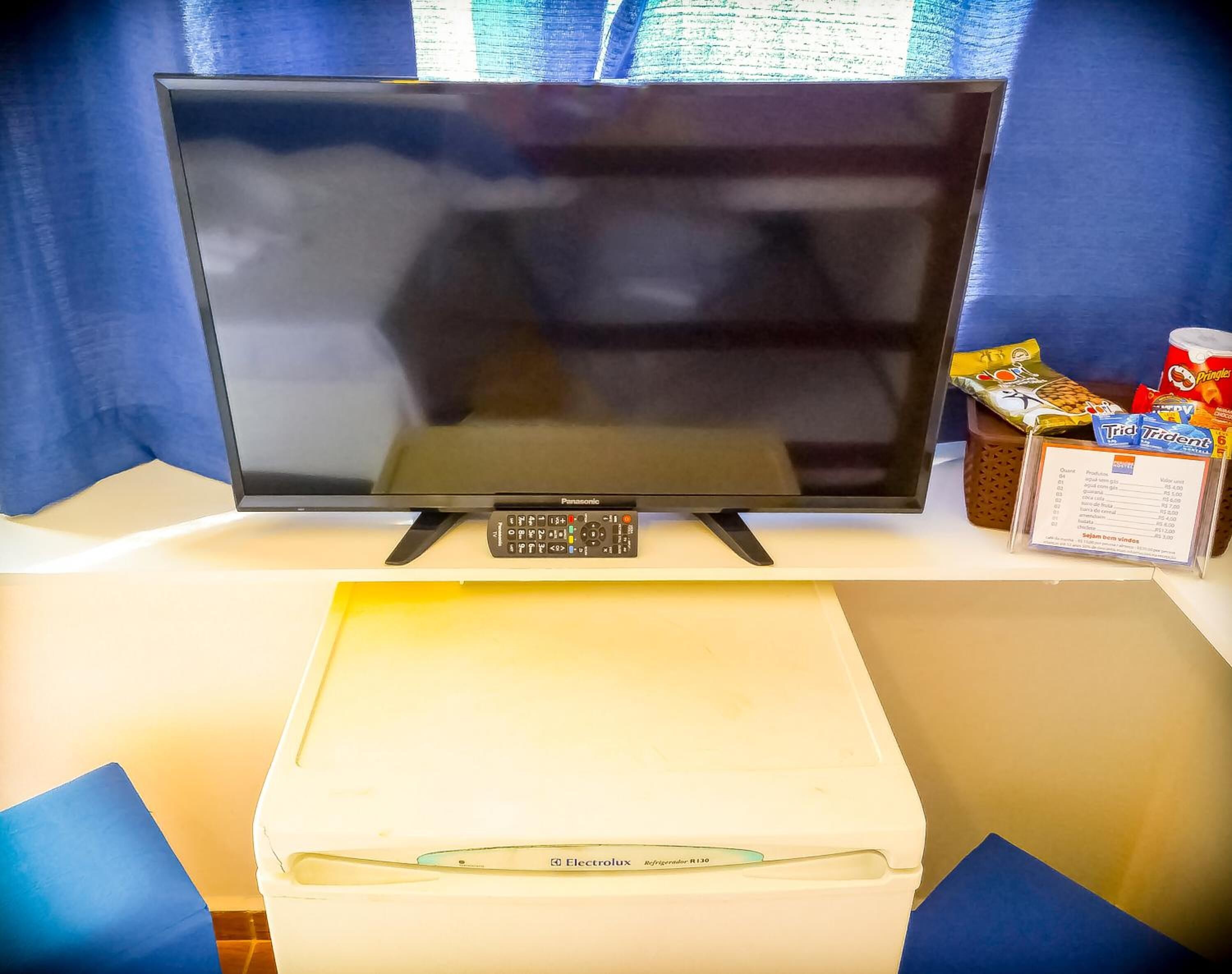 Un téléviseur Panasonic posé sur une table blanche, avec une télécommande à proximité. En dessous, une boîte portant l’inscription « Electrolux », et à droite, des collations et des boissons.