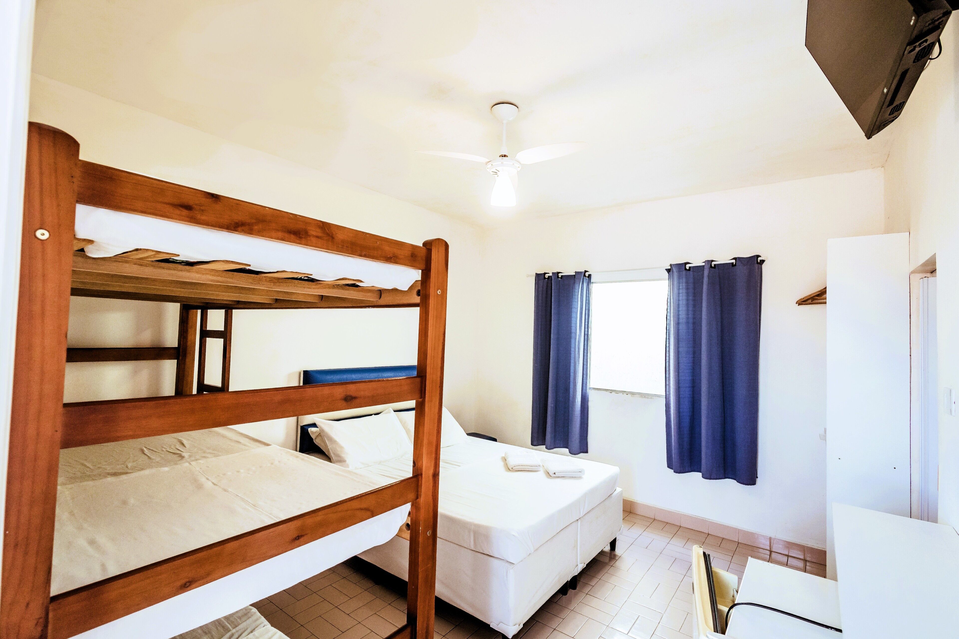Una cama individual con dosel y estructura de madera, una cama nido con ropa blanca, un ventilador de techo, una ventana con cortinas azules y un refrigerador blanco.