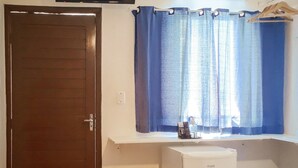 Une petite pièce avec une porte, un petit réfrigérateur blanc, un coussin bleu, une fenêtre avec des rideaux bleus et un téléviseur fixé au mur.