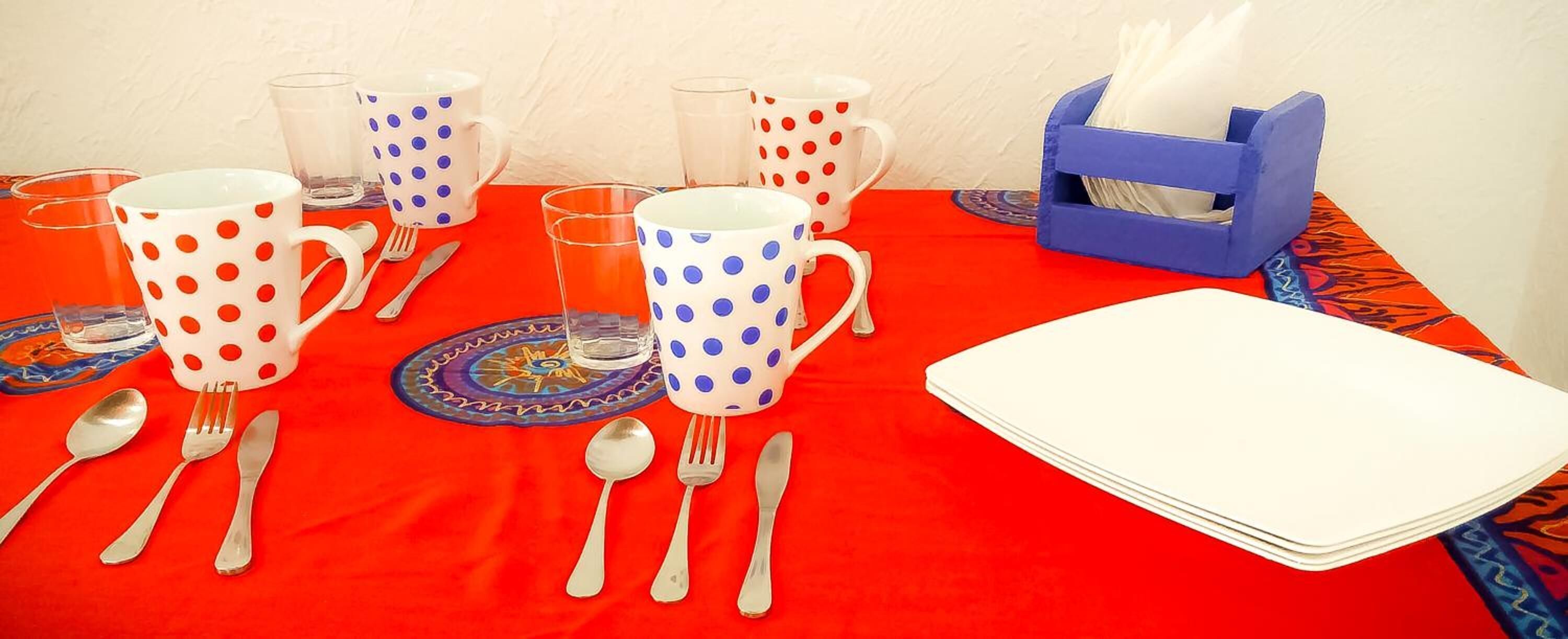 Ein Tisch ist mit weißen Tellern, blau-rot gepunkteten Tassen und silbernem Besteck auf einer roten Tischdecke gedeckt.