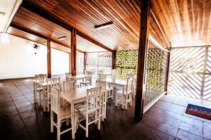 Un espacio de comedor al aire libre cubierto con mesas y sillas de madera, piso de baldosas y vista al exterior a través de una valla decorativa.