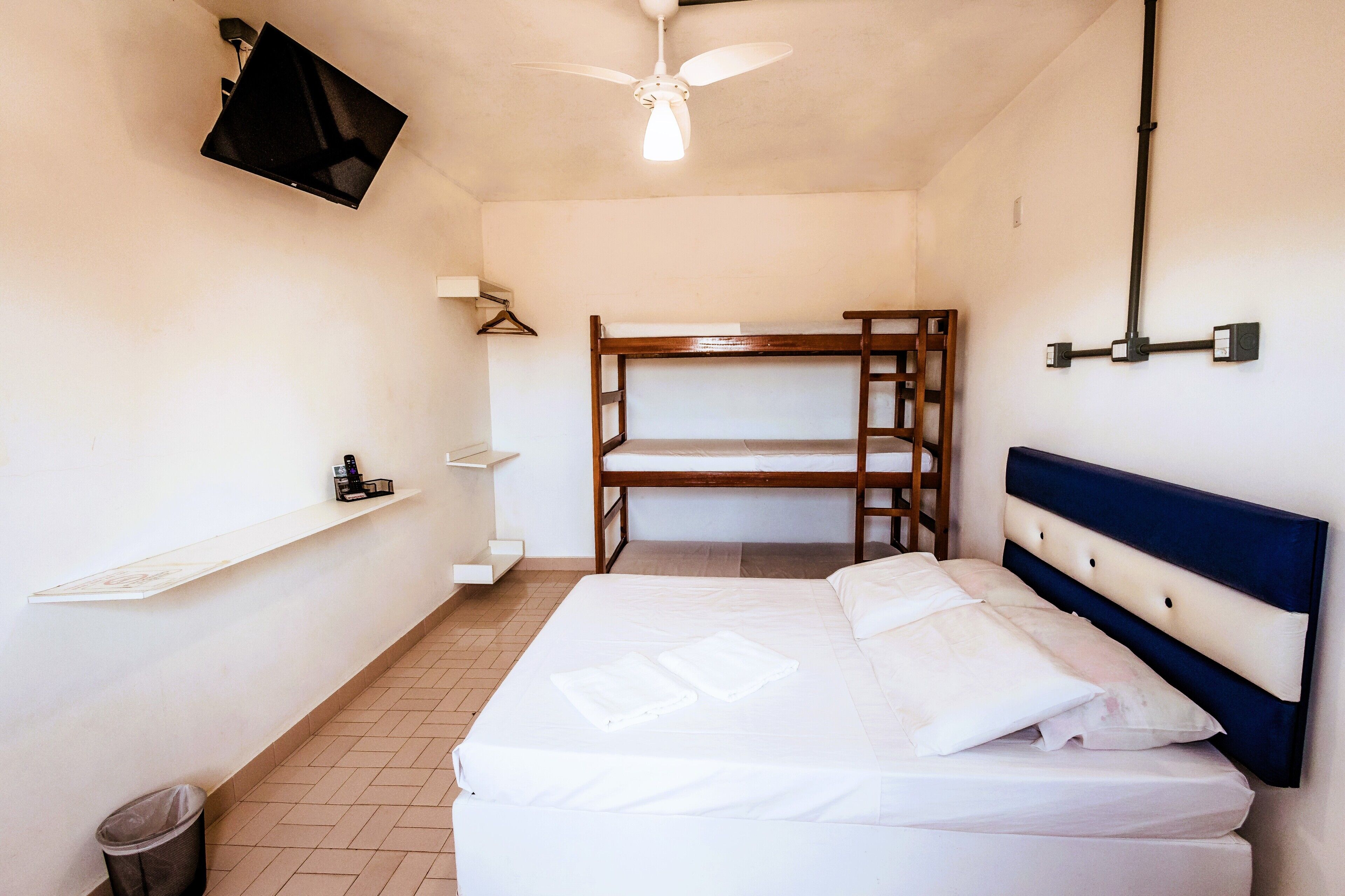 A bedroom with a bunk bed, a single bed, a TV mounted on the wall, a ceiling fan, and a small trash bin.
