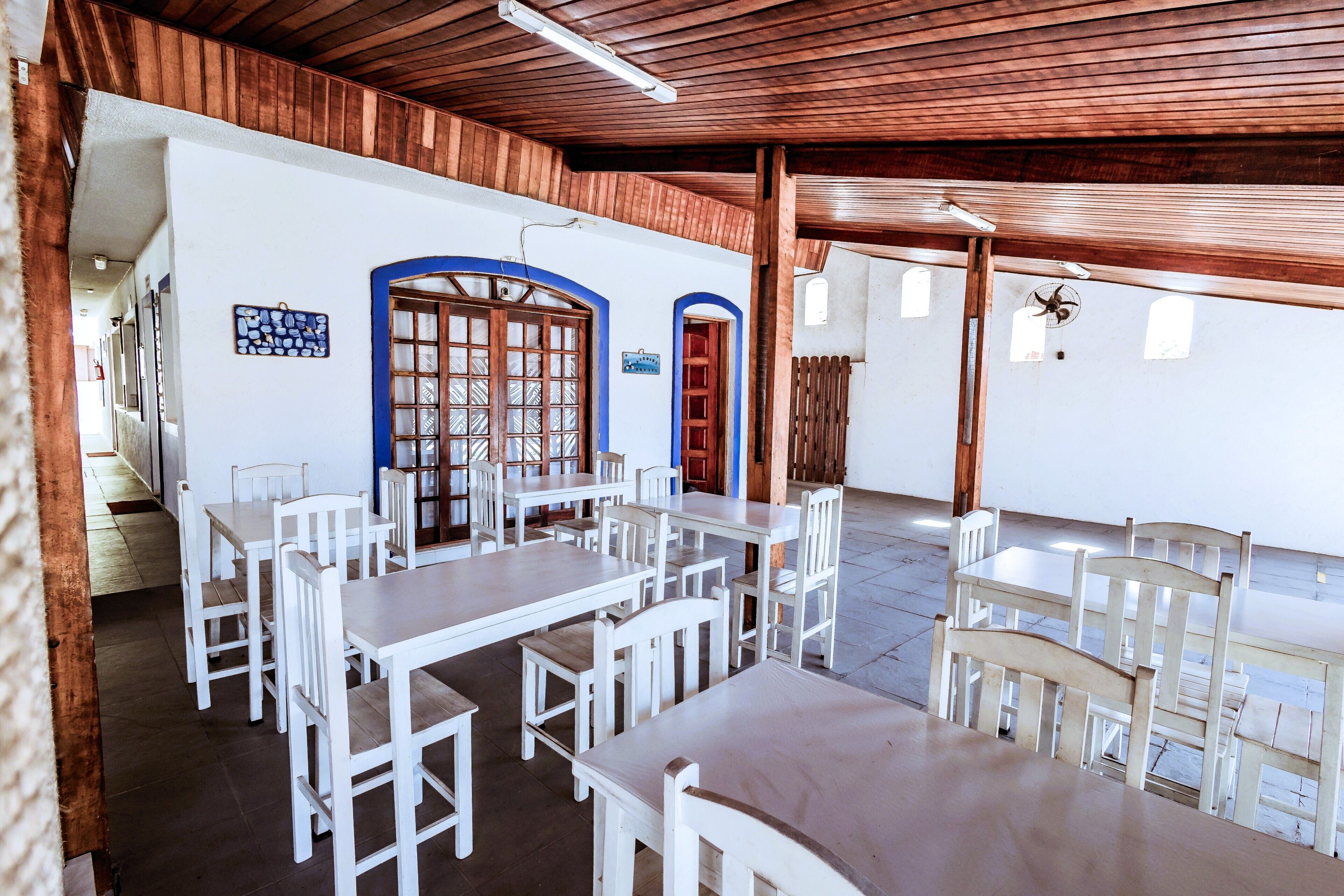 Un espace repas avec des tables et des chaises blanches, des poutres en bois au plafond et une fenêtre à cadre bleu.