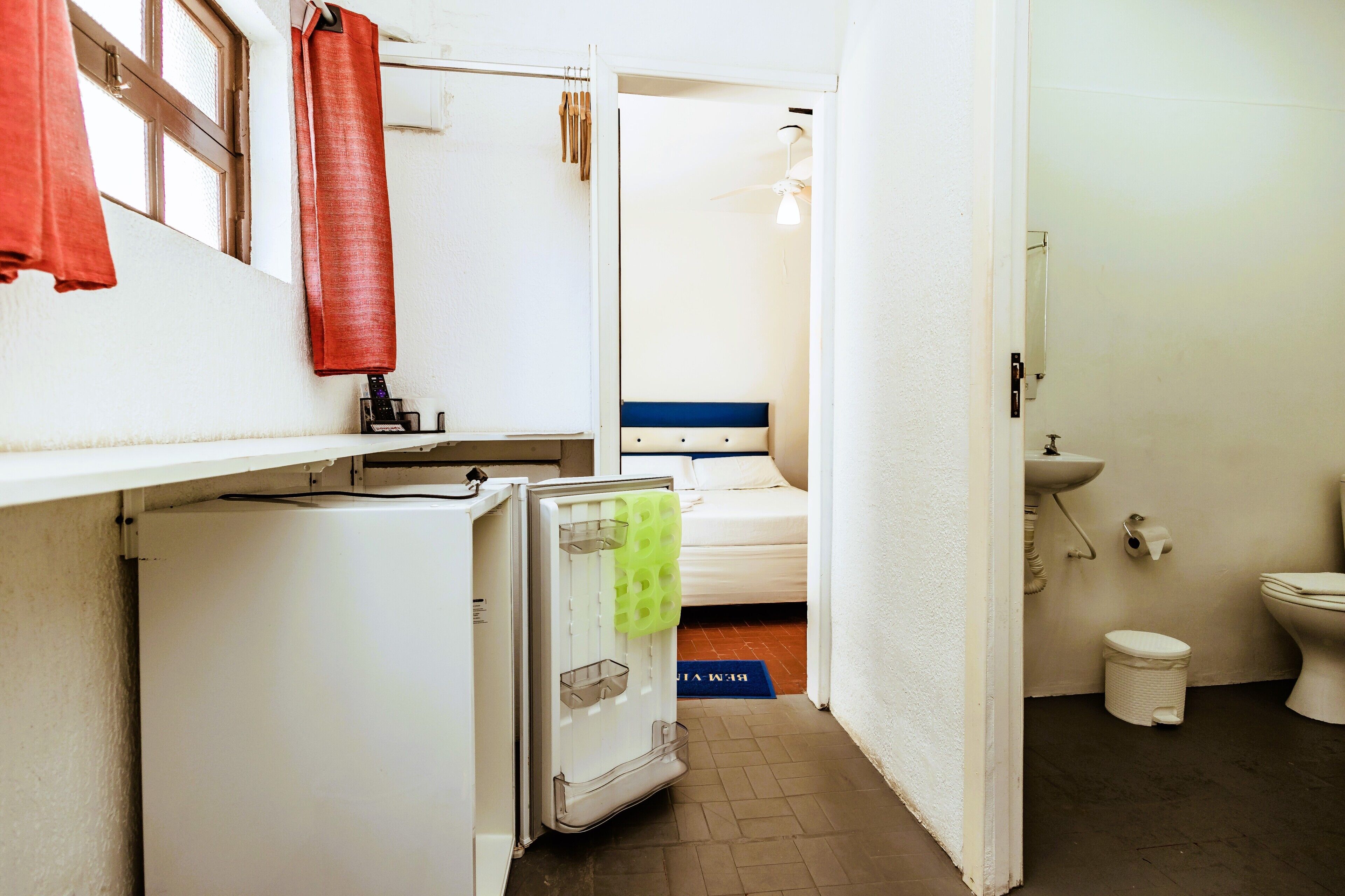 A compact hotel room with a bed, a small kitchenette, a bathroom, and a window with a red curtain.