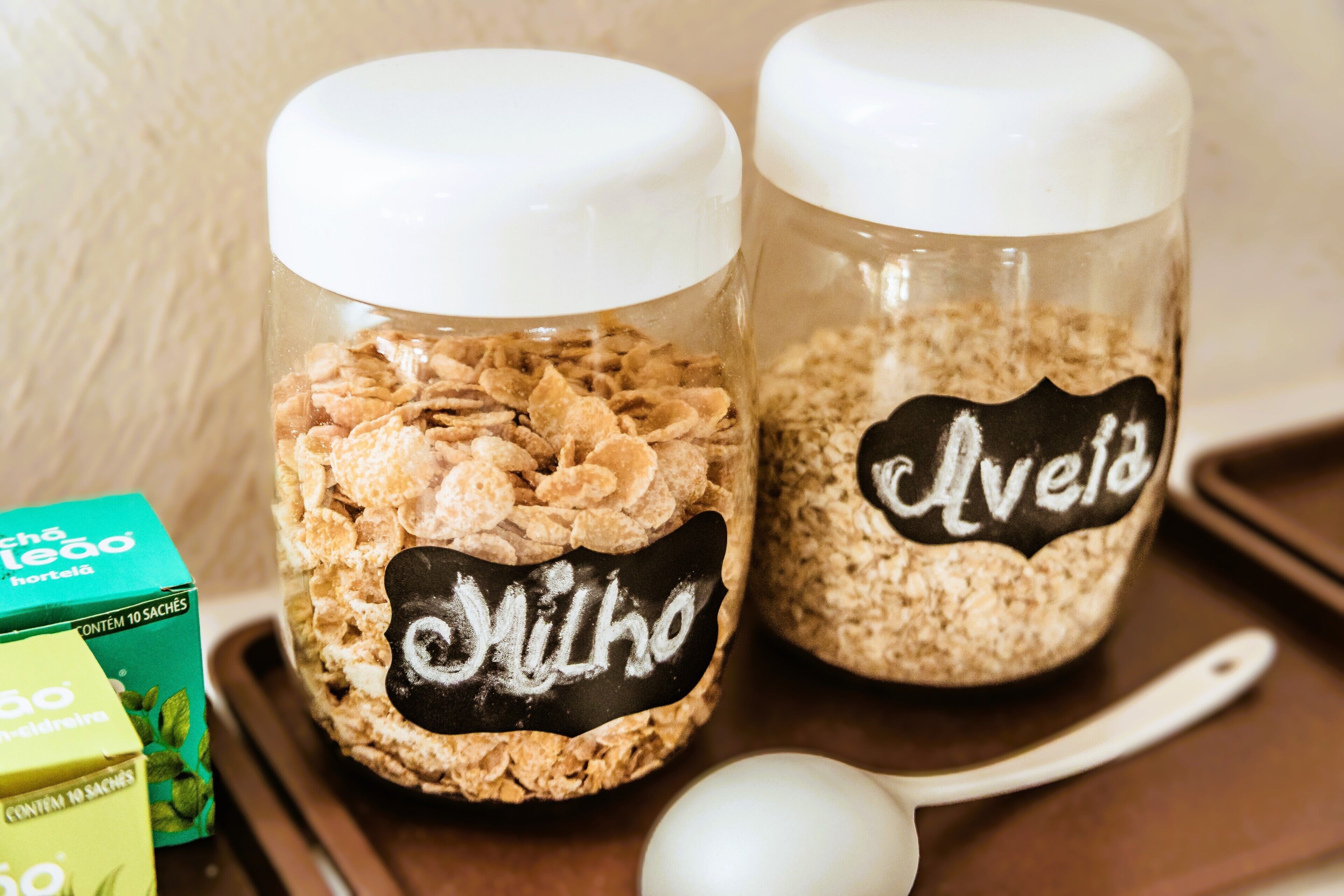 Two glass jars with white lids, one labeled 'Avelho' and the other 'Hortelã,' containing different grains.