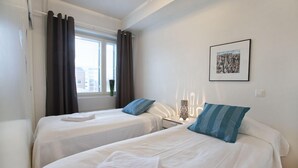 A hotel room with two beds, a window with curtains, a bedside table with a lamp, and a framed picture on the wall. - RovaVisit Center (Rovaniemi)