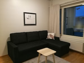 Ein schwarzes Sofa mit Kissen, ein kleiner Holzschemel, ein gerahmtes Bild an der Wand und ein Fenster mit Vorhängen.
