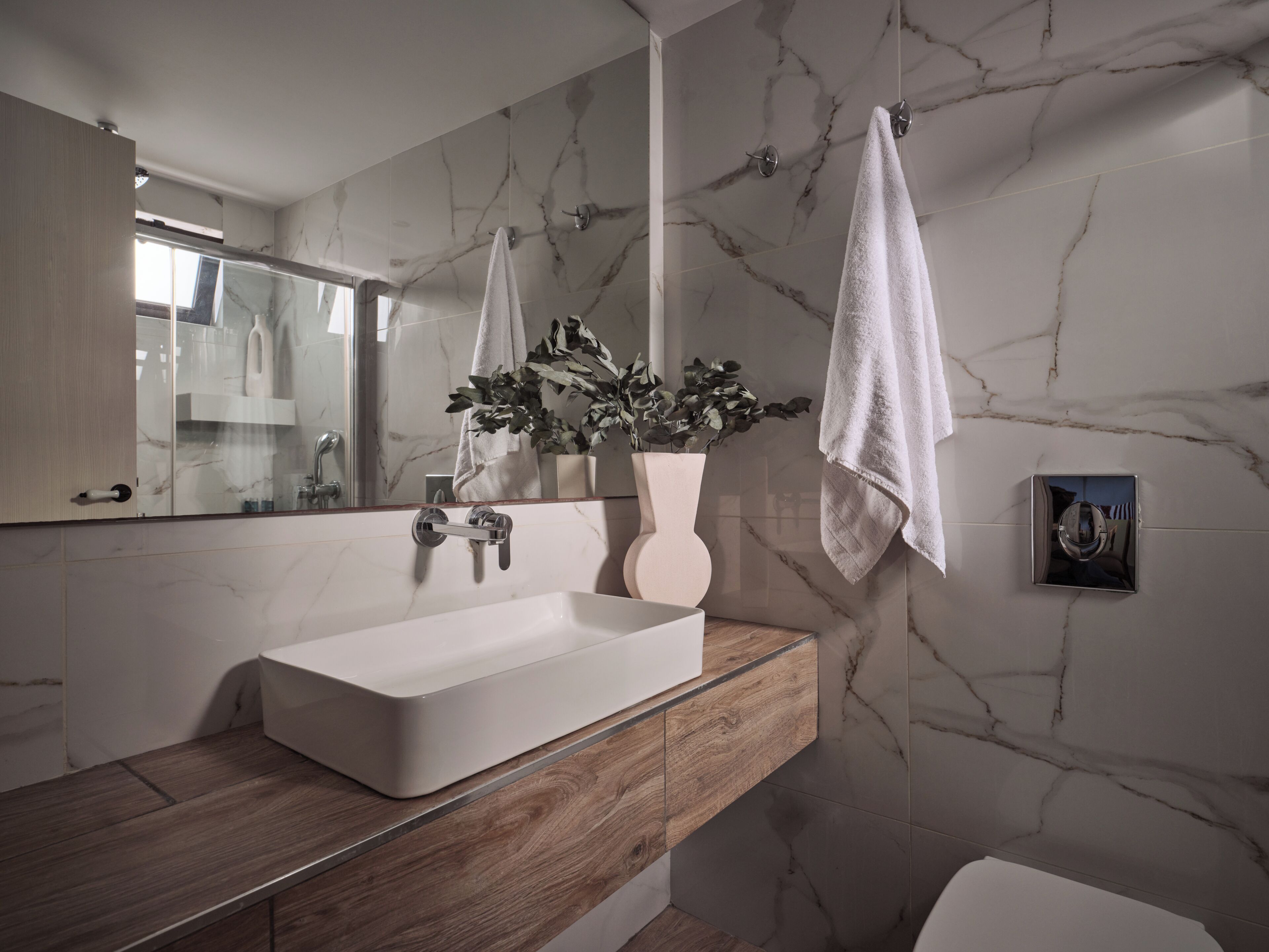 Un baño moderno con pared de mármol, tocador de madera, lavamanos blanco y una planta en maceta.