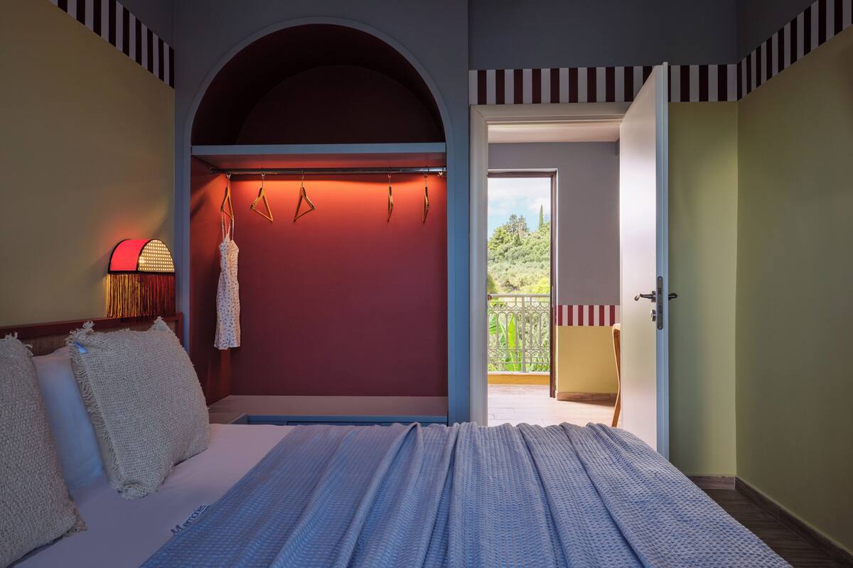 A bedroom with a bed, a hanging light fixture, a view through an open door, and a balcony.