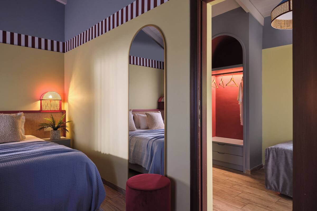 A hotel room with two beds, a mirror, a red stool, and a wardrobe with a red interior.