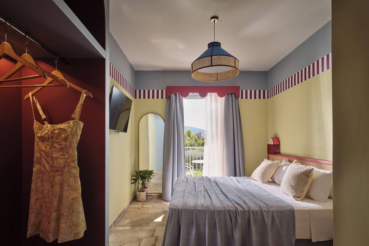A bedroom with a bed, a hanging dress, a potted plant, and a view through a window.