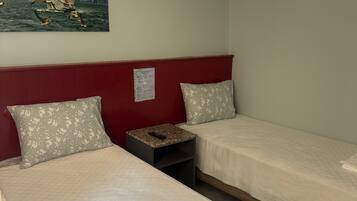 A hotel room with two beds, a red headboard, floral pillows, a nightstand with a remote control, and a painting of a boat on the wall.