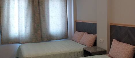 Quarto de hotel com duas camas, cada uma com cabeceira e almofadas, uma mesinha e uma janela com cortinas.