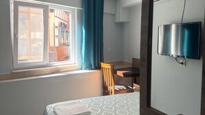 A bedroom with a bed, a desk, a chair, a TV, and a window with curtains. - Kisla Pansiyon (Antalya)