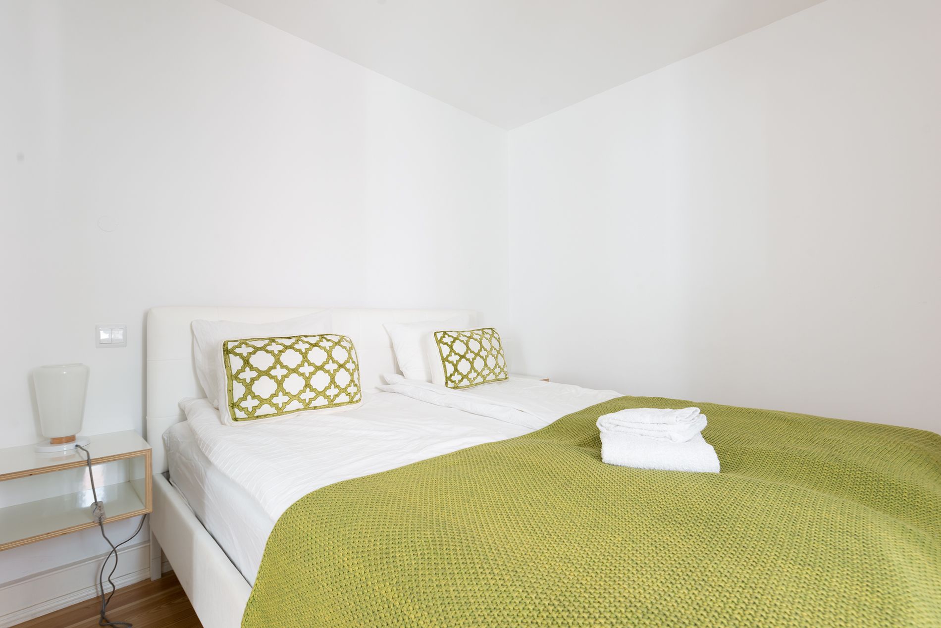 Un dormitorio minimalista con una cama blanca, dos almohadas estampadas, una manta verde y una mesita de noche con una lámpara.