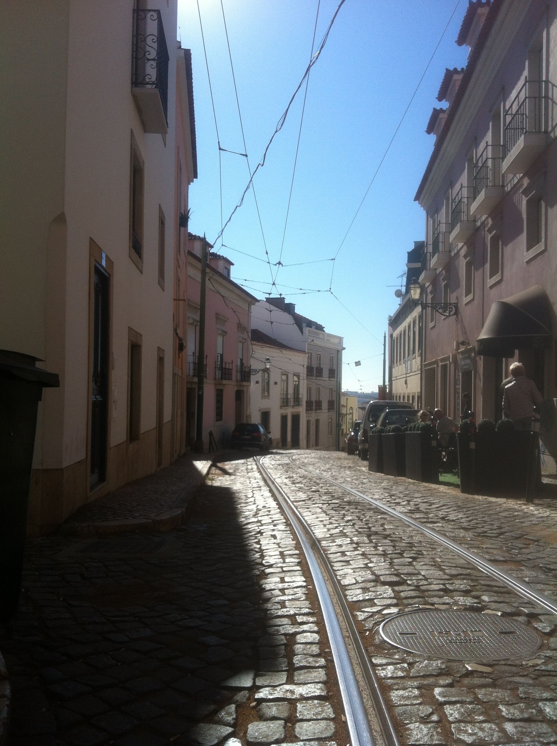 A cobblestone street with tram tracks, buildings on either side, and overhead tram lines.