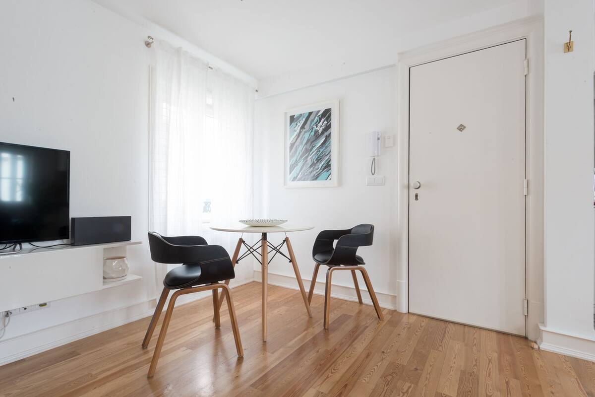 A modern living room with a flat-screen TV, two black chairs, a small round table, and a door.