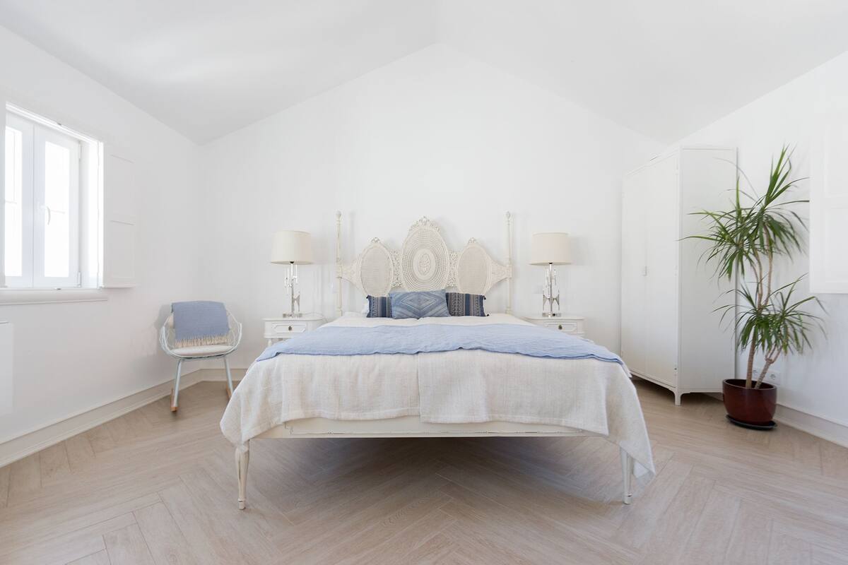 A bedroom with a bed, bedside lamps, a chair, a potted plant, and a window.