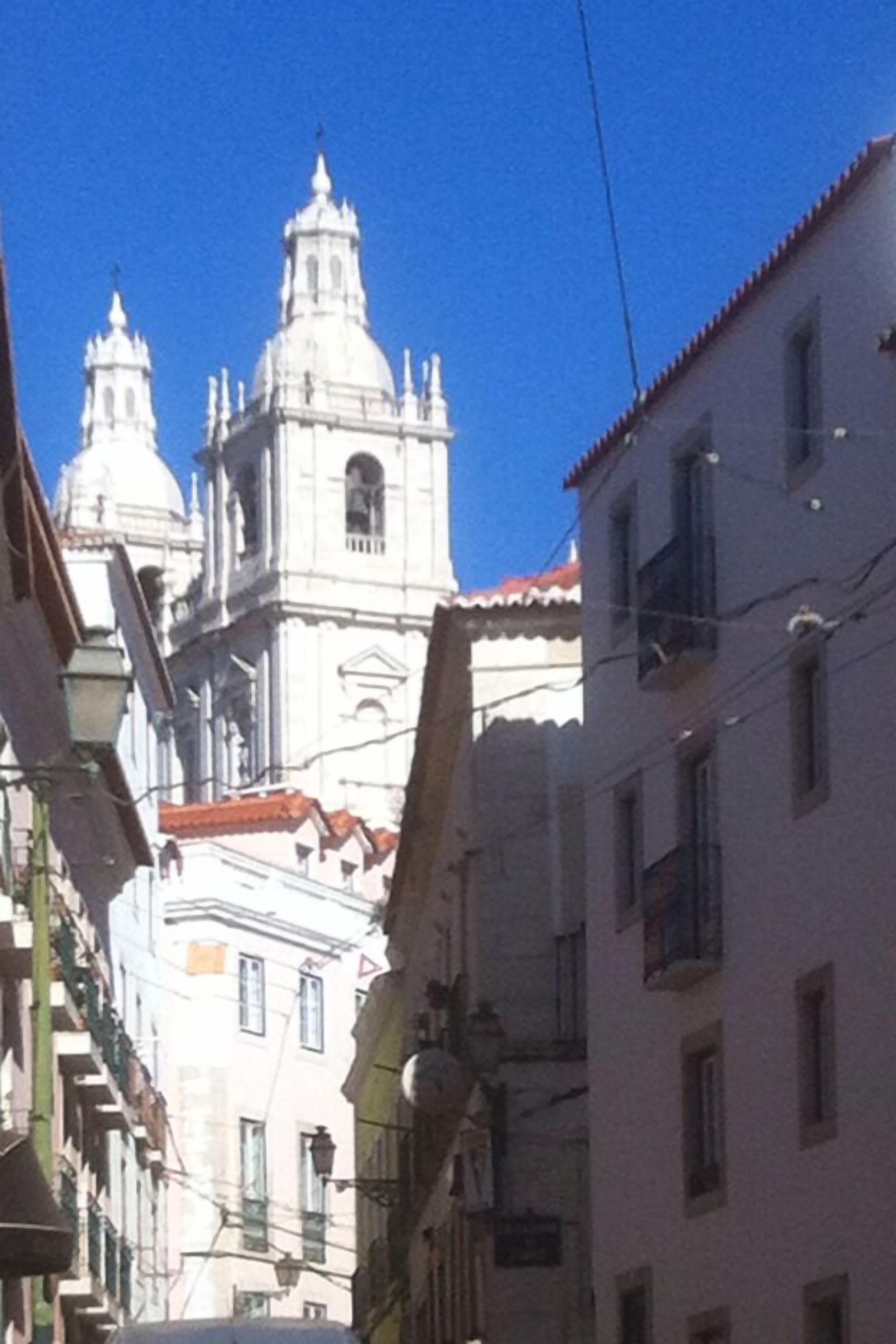 Vista de una calle con una iglesia blanca destacada, con dos cúpulas y un campanario, rodeada de edificios con balcones y tejados de tejas rojas.
