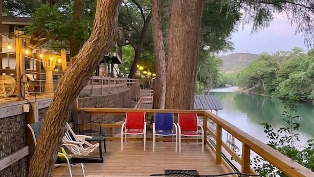 A wooden deck with chairs overlooking a river, surrounded by trees and a stone wall. - Ponderosa Cabin (New Braunfels)