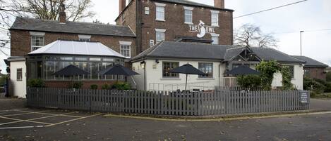 A pub named "The Owl" with a white facade, brick chimney, and a fenced outdoor seating area with umbrellas.