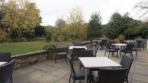 An outdoor patio with wicker tables and chairs, surrounded by a stone wall and trees. - Owl, Hambleton by Marston's Inns (Selby)