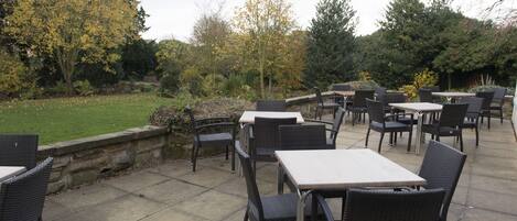 An outdoor patio with wicker tables and chairs, surrounded by a stone wall and trees.