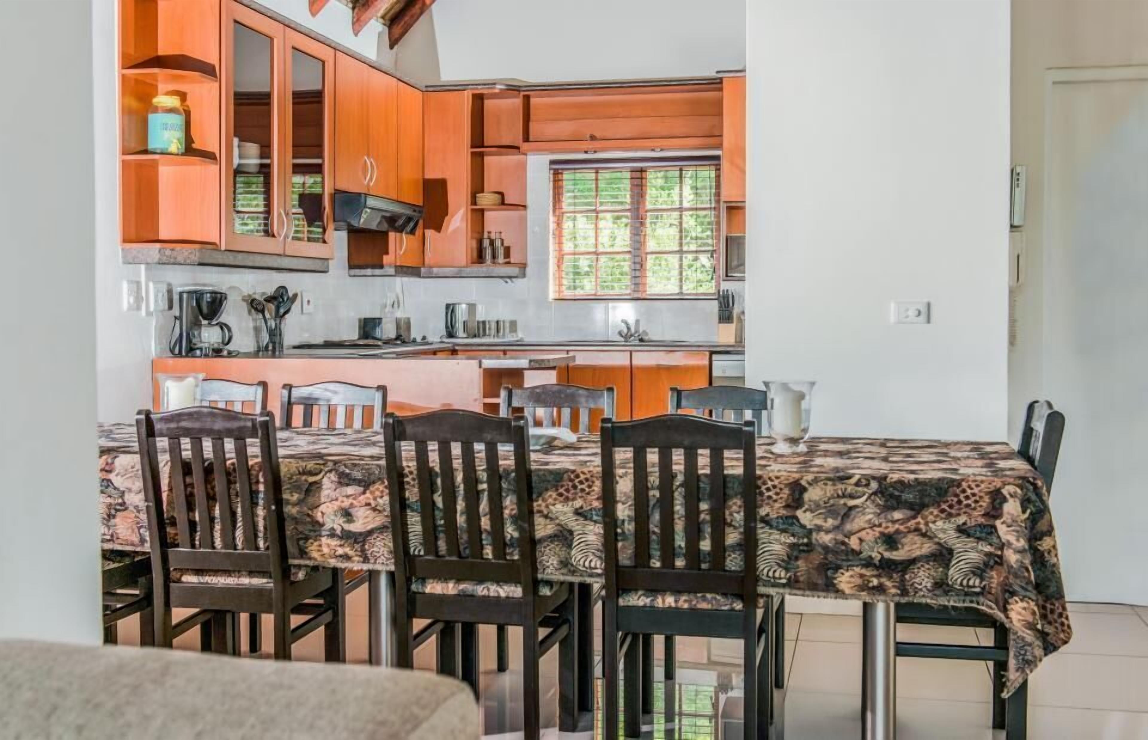 A compact kitchen with wooden cabinets, a dining table with chairs, and a window with a view of greenery.