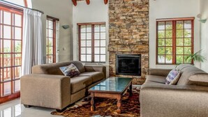 A living room with two sofas, a coffee table, a stone fireplace, and large windows with shutters. - St Francis Bay Luxury Lodge 2 (Saint Francis Bay)