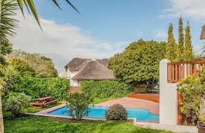 A pool surrounded by lush greenery, a thatched-roof house, and a wooden fence.
