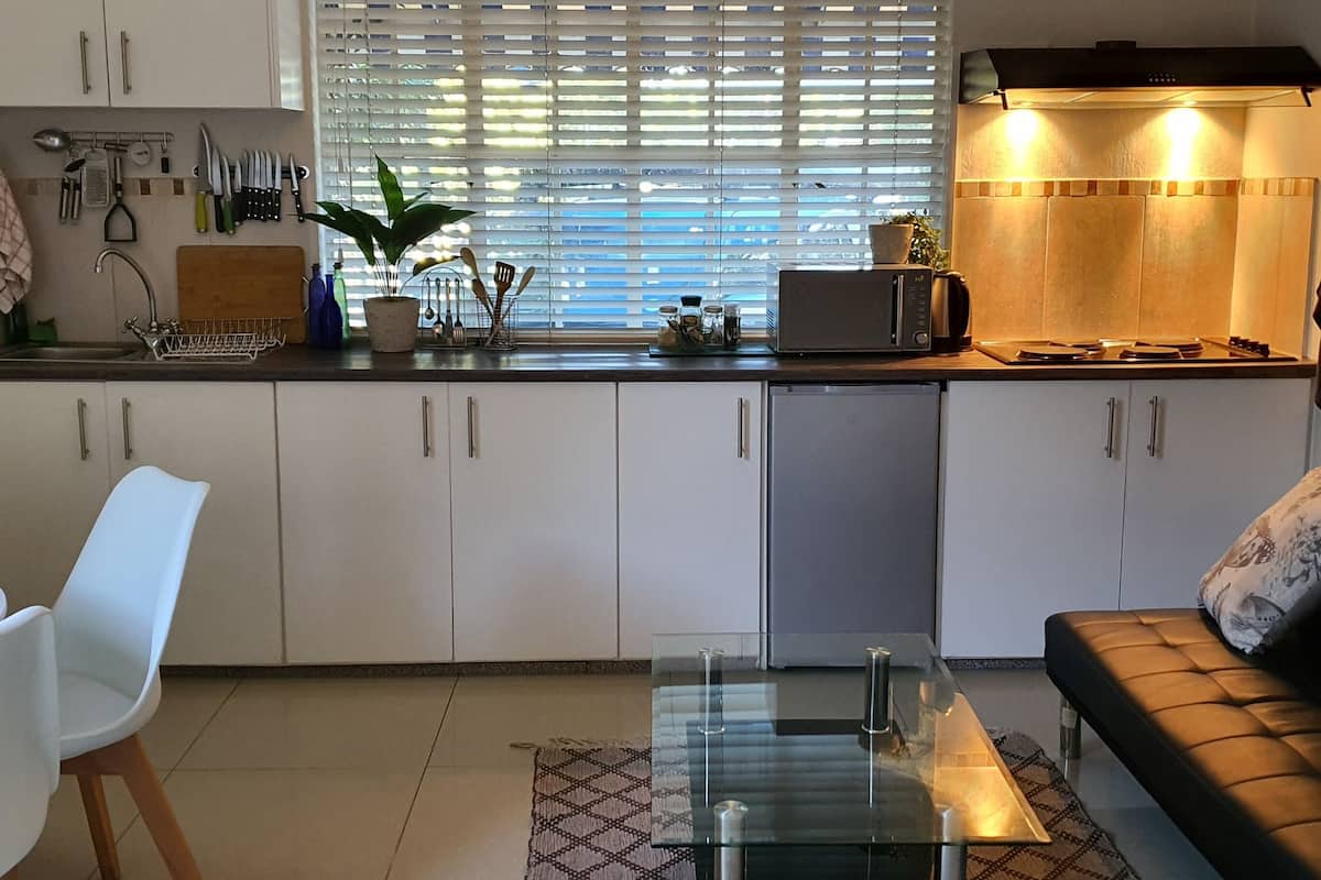 A compact kitchen with white cabinets, a dining area with a white table and chairs, and a black leather sofa with patterned cushions.