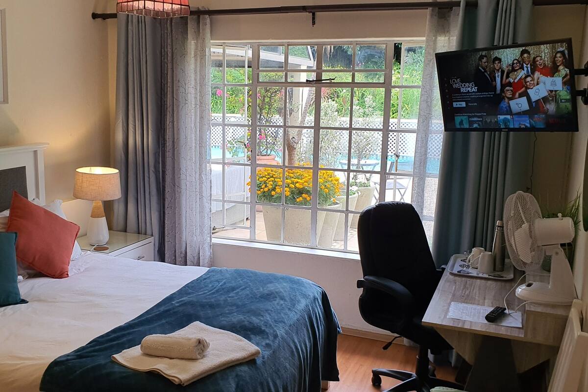 A hotel room with a bed, a desk with a chair, a TV, and a window with a view of greenery and flowers.