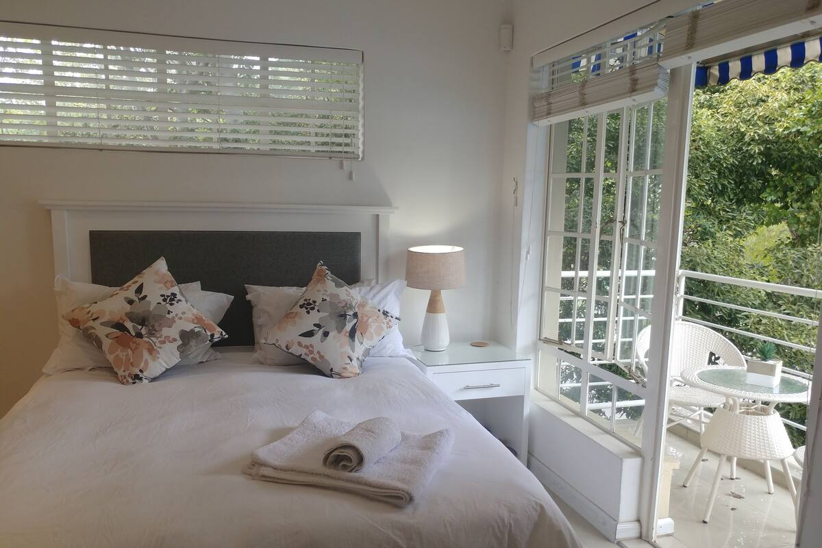 A hotel room with a bed, pillows, a nightstand, a lamp, and a balcony with a table and chairs.