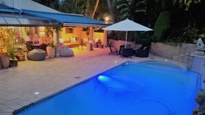 A swimming pool with a blue tint, surrounded by a patio area with outdoor furniture and a covered seating area. - Pepper Tree BnB (Sandton)