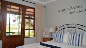 A bedroom with a bed, a nightstand, a wall with a motivational quote, a window with curtains, and a view of greenery outside. - African Sun Guest House (George)