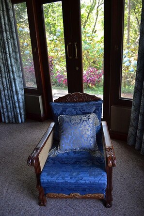 Un sillón azul con un cojín estampado en una habitación con ventanas grandes, cortinas y vista a zonas verdes.