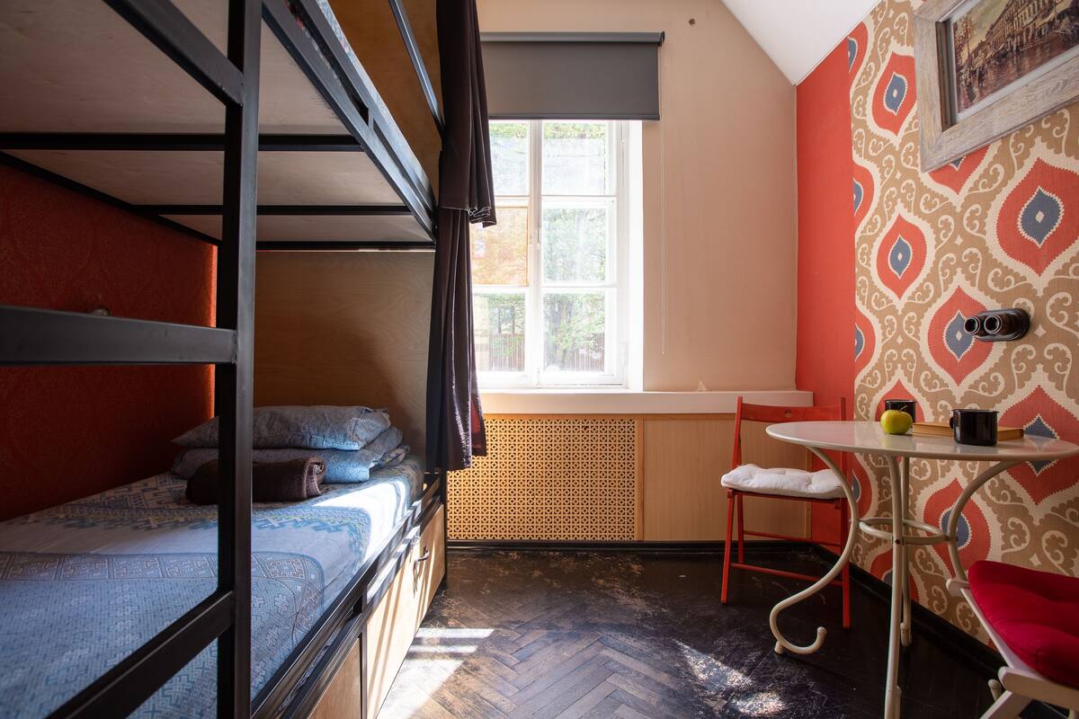 Ein Schlafsaalzimmer mit Etagenbetten, einem kleinen Tisch mit Stuhl, einem Heizkörper und einem Fenster mit Jalousien.