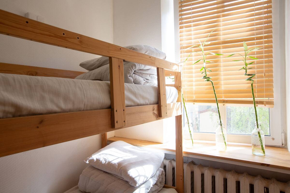 Ein Stockbett mit Holzrahmen, ein Fenster mit Rollo und zwei Flaschen mit Pflanzen auf dem Sims.