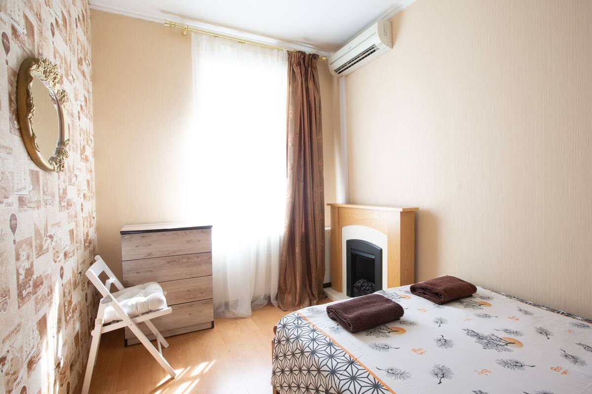 Ein Schlafzimmer mit einem Bett, einer hölzernen Kommode, einem Sessel, einem Kamin und einem Spiegel an der Wand.