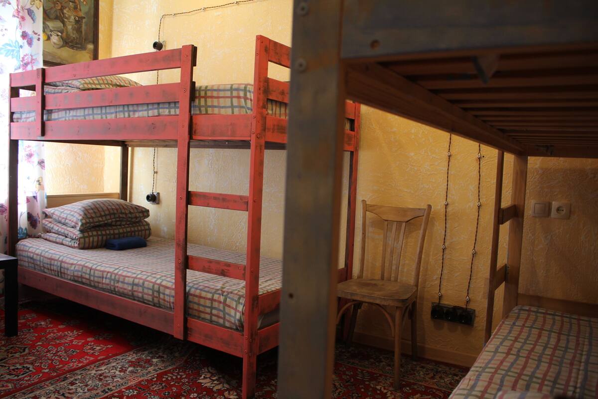 Ein Etagenbett mit Karo-Bettwäsche, ein Holzstuhl und ein gemustertes Teppich in einem Raum mit Holzboden und -wänden.