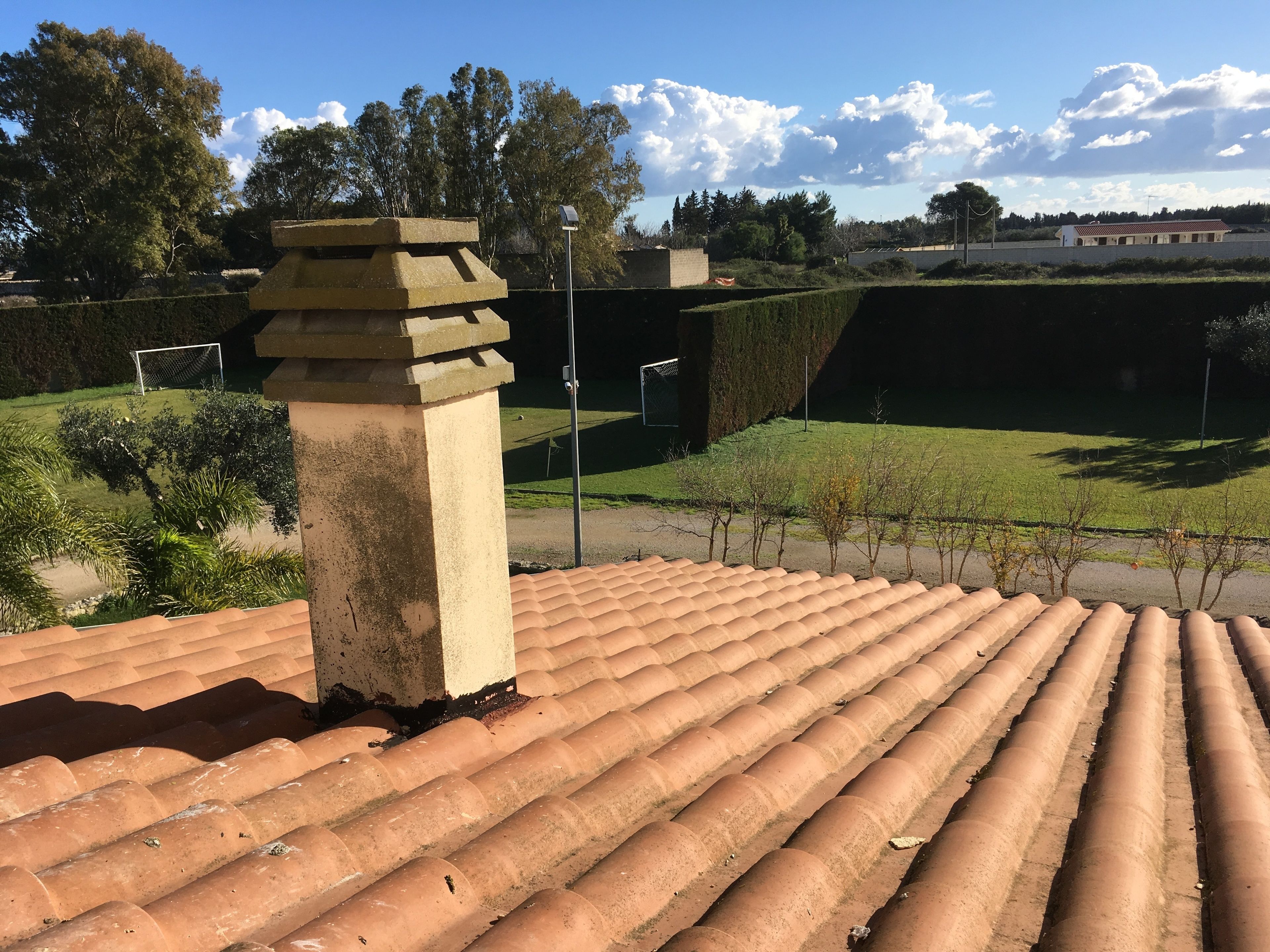 Eine Dachterrasse mit Terrakottafliesen, einem Steinsschornstein und Blick auf ein Sportfeld und Bäume.