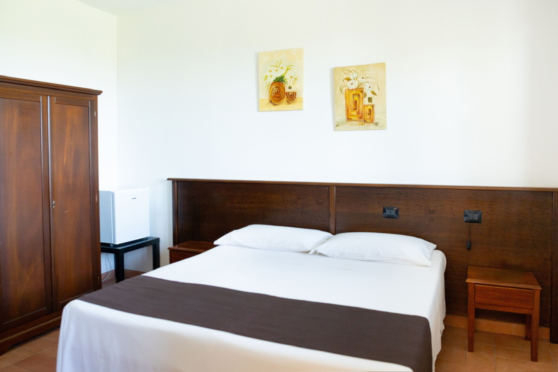 A hotel room with a large bed, wooden headboard, two bedside tables, and a wardrobe.