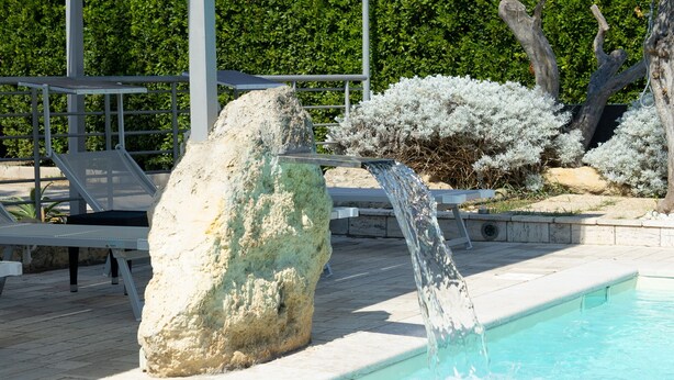 A swimming pool with a waterfall feature, surrounded by lounge chairs and a stone structure. - Giardini di Marzo Lecce B&B (Lecce)