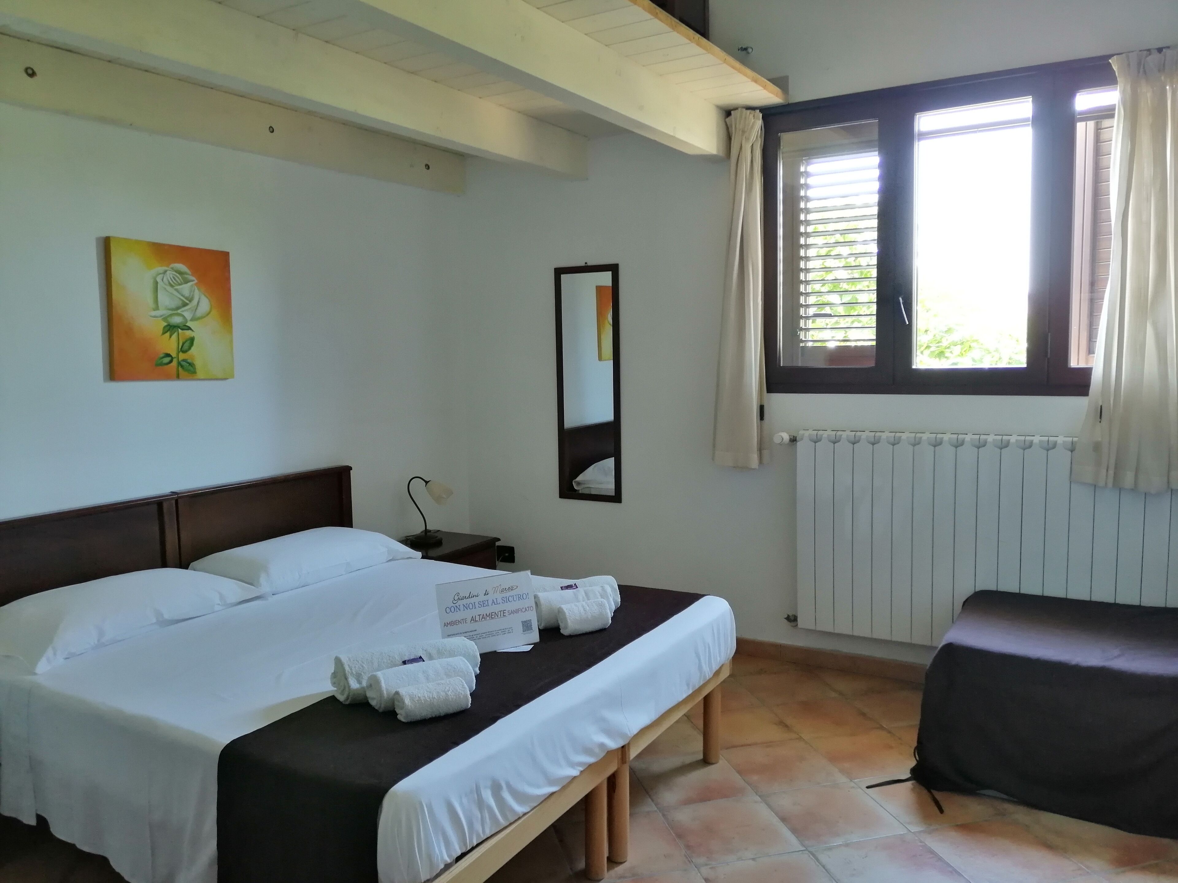 Habitació d'hotel amb llit, una banca, una tauleta de nit, làmpada, quadre, finestra amb cortines i un radiador.