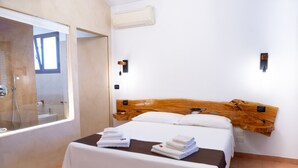 A hotel room with a wooden headboard, a bed with white linens, a small table with folded towels, a window with a view of the outside, and a wall-mounted air conditioner. - Giardini di Marzo Lecce B&B (Lecce)