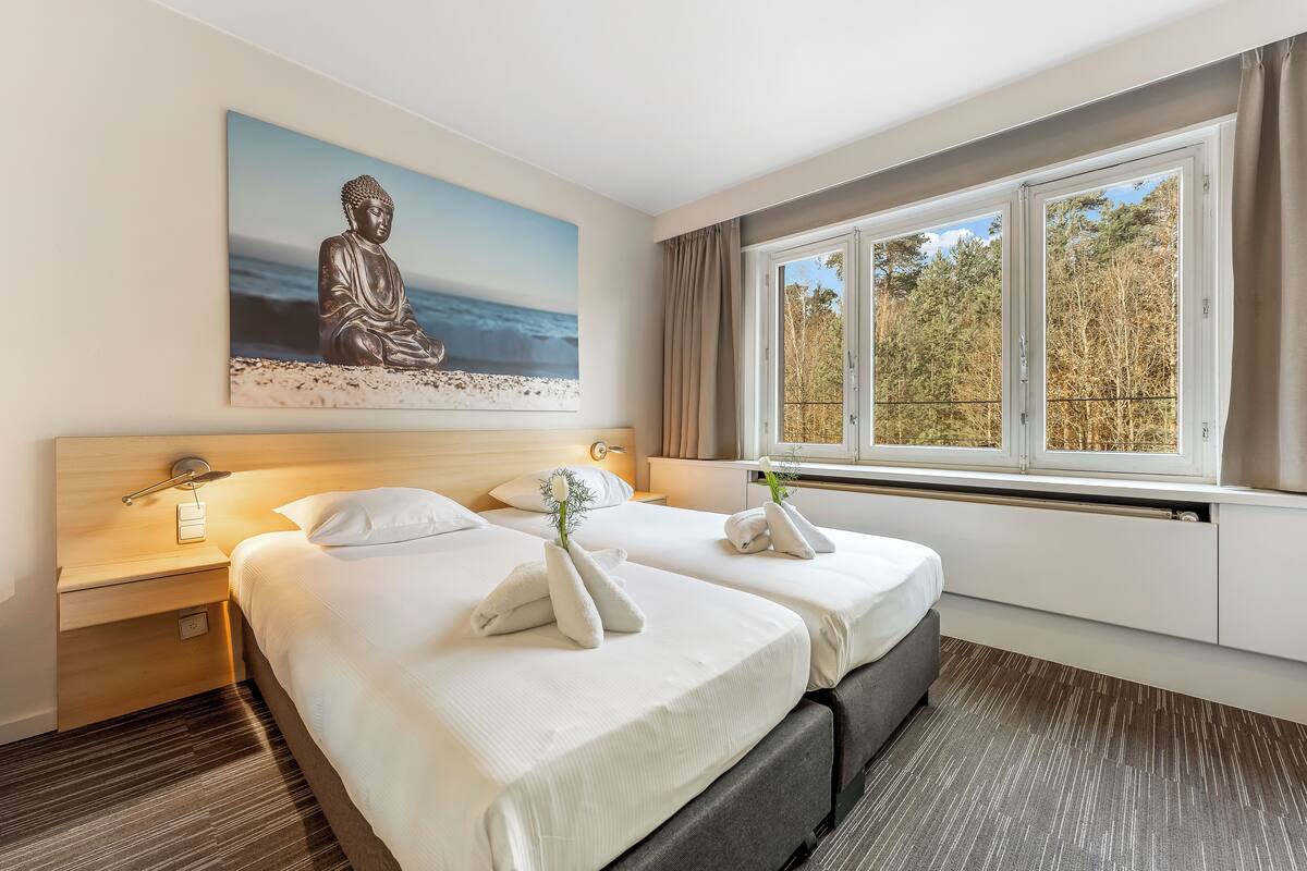 A hotel room with two beds, a large Buddha painting, a window with a view of trees, and a bedside table with a lamp.