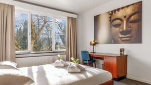 A hotel room with a bed, a desk, a chair, a window with curtains, and a Buddha painting on the wall. - Domein Martinus (Zoersel)