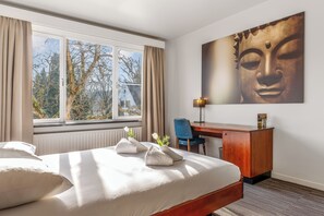 A hotel room with a bed, a desk, a chair, a window with curtains, and a Buddha painting on the wall. - Domein Martinus (Zoersel)