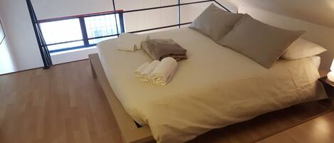 Un dormitorio moderno con piso de madera, una cama con ropa blanca y dos almohadas, y una ventana con vista al cielo.