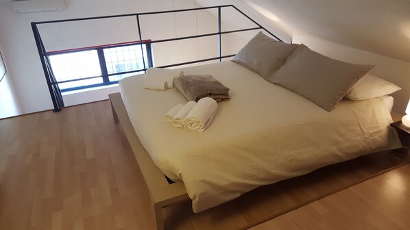 A modern bedroom with a wooden floor, a bed with white bedding and two pillows, and a window with a view of the sky.